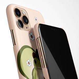 May include: A pink phone case with a green frog wearing a cowboy hat and white flowers. The case is designed for an iPhone with a triple camera lens.
