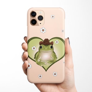 May include: A pink iPhone case with a green frog wearing a brown cowboy hat inside a heart. The case is decorated with white flowers.