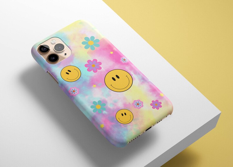 May include: A colorful tie-dye phone case with a pink, blue, and yellow background. The case features a variety of yellow smiley faces and pink and blue flowers.