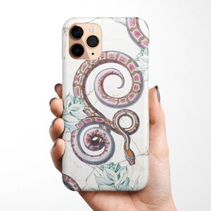 Może przedstawiać: Białe etui na iPhone z wzorem różowego i brązowego węża. Wąż jest zwinięty w spiralę i otoczony białymi kwiatami.