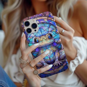 May include: A colorful phone case with a stained glass design of a frog. The case is blue, purple, and green with a white outline. The frog has large, round eyes and a detailed design.