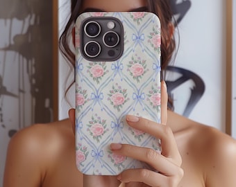 May include: A white iPhone case with a floral pattern featuring pink roses and blue bows. The case has a camera lens cutout and a white background.