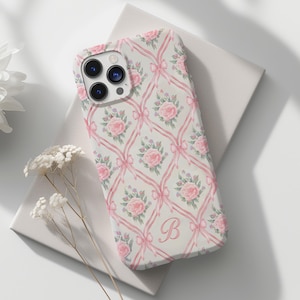 May include: A white phone case with a pink floral pattern and a pink letter "B" in the center. The case has a camera lens cutout and a cutout for the phone's buttons.
