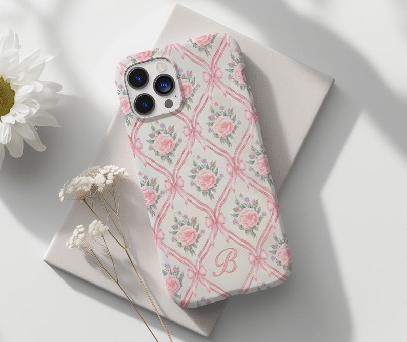 May include: A white phone case with a pink floral pattern and a pink letter "B" in the center. The case has a camera lens cutout and a cutout for the phone's buttons.