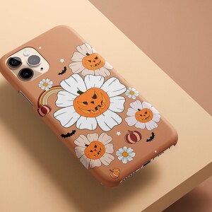 Pu&ograve; includere: Una custodia per iPhone marrone a tema Halloween. La custodia presenta fiori bianchi con zucche arancioni e pipistrelli.