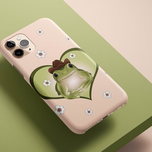 May include: A pink iPhone case with a green frog wearing a brown cowboy hat in a green heart. The case is decorated with white flowers.