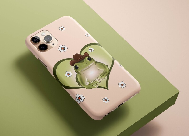 May include: A pink iPhone case with a green frog wearing a brown cowboy hat in a green heart. The case is decorated with white flowers.