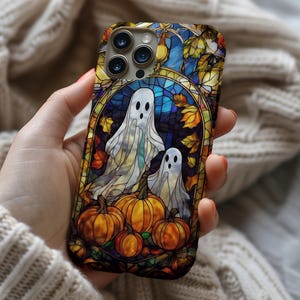 Puede incluir: Una funda de teléfono colorida con un diseño de vidriera que presenta dos fantasmas blancos, calabazas y hojas de otoño. La funda tiene un fondo azul oscuro y un borde dorado, con temática de Halloween.