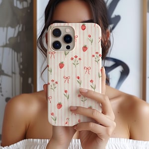 May include: A phone case featuring a light pink and white vertical striped background with a repeating pattern of small red strawberries, tiny red flowers on green stems, and delicate red bows scattered across the entire surface area.