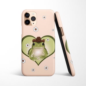 May include: A pink phone case with a green frog wearing a cowboy hat inside a heart. The frog is surrounded by white flowers.