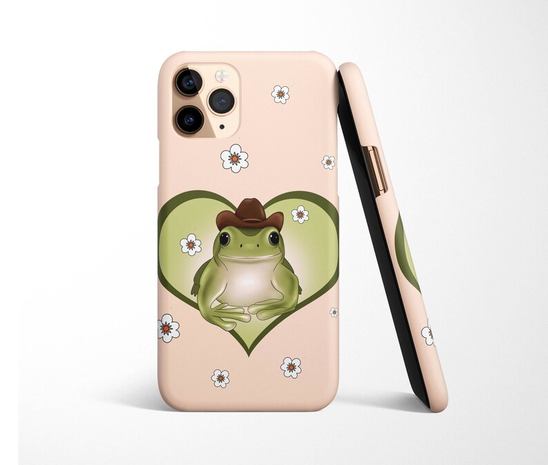 May include: A pink phone case with a green frog wearing a cowboy hat inside a heart. The frog is surrounded by white flowers.
