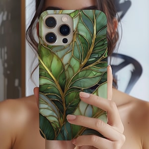 Puede incluir: Una funda de teléfono con un diseño de hojas verdes y doradas. La funda tiene un acabado brillante y un patrón detallado de hojas con detalles dorados. La funda del teléfono está en la mano de una persona.