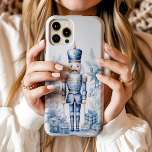 Op de afbeelding: Een witte telefoonhoes met een blauwe en witte aquarel-illustratie van een notenkraker die een zwaard vasthoudt en voor een blauw en wit bloemenontwerp staat. De notenkraker draagt een blauw en gouden uniform met een rode en witte hoed.