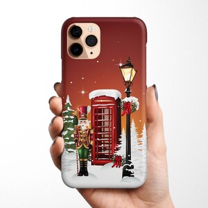 May include: A red phone booth with a snow-covered roof stands next to a street lamp with a lit lantern. A nutcracker in a green and gold uniform stands in front of the phone booth. The background is a red and brown gradient with white snowflakes.