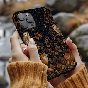 Peut inclure: Une coque de smartphone noire ornée d'un imprimé détaillé représentant deux hiboux, deux renards et un croissant de lune au milieu d'une forêt dense de feuilles d'automne, de fleurs dorées et de feuillage sombre, avec un triple objectif photo.