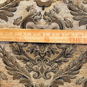 Puede incluir: Primer plano de una tela con un patr&oacute;n de damasco detallado en tonos marr&oacute;n, dorado y negro. Una regla de madera, de unos 30 cm de largo, descansa sobre la tela. La tela tiene una textura tejida y un dise&ntilde;o cl&aacute;sico y ornamentado.