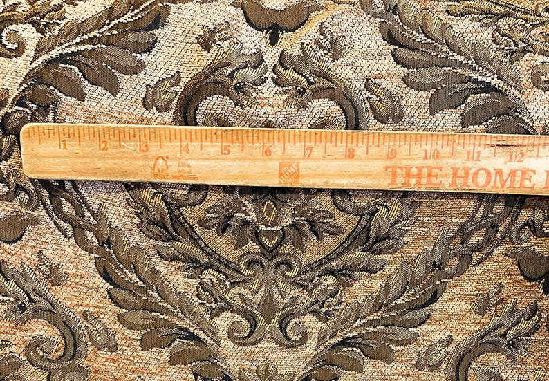 Puede incluir: Primer plano de una tela con un patr&oacute;n de damasco detallado en tonos marr&oacute;n, dorado y negro. Una regla de madera, de unos 30 cm de largo, descansa sobre la tela. La tela tiene una textura tejida y un dise&ntilde;o cl&aacute;sico y ornamentado.