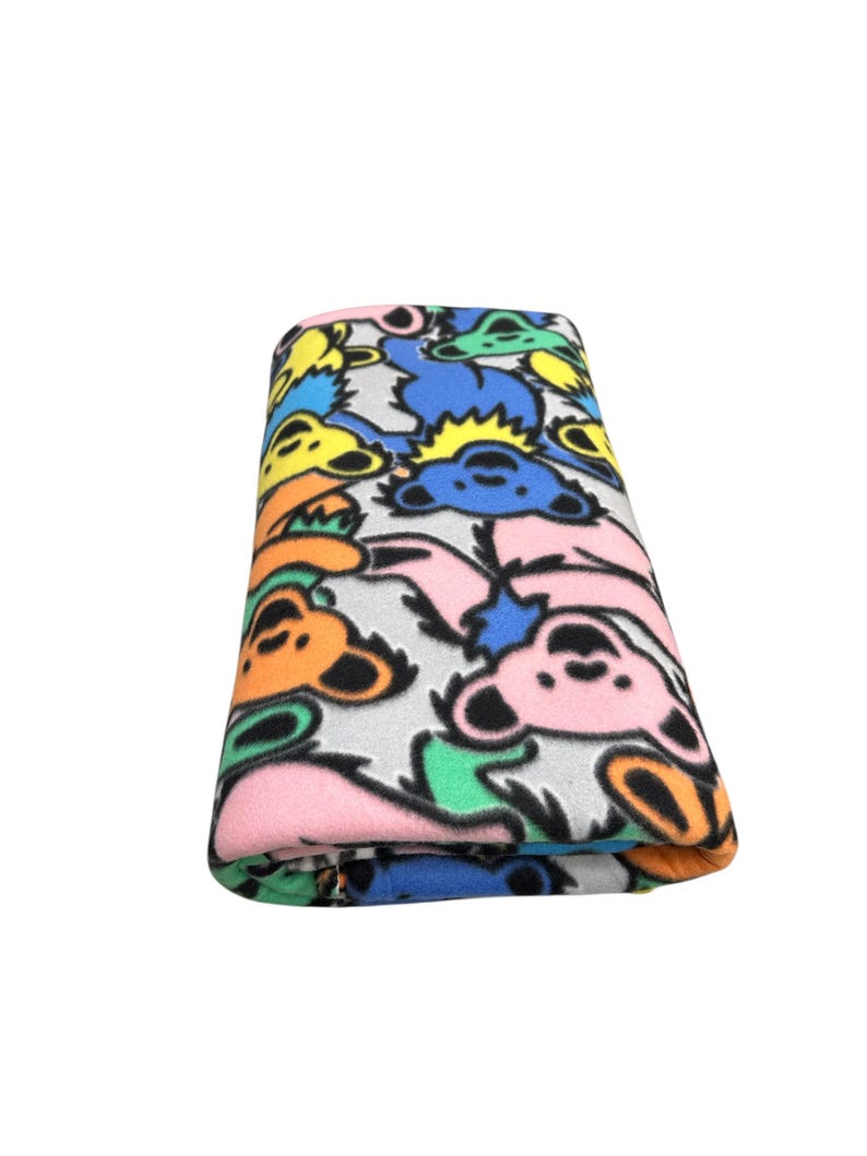 Puede incluir: Manta de forro polar plegada con un patr&oacute;n repetido de ilustraciones de osos coloridos en tonos rosa, naranja, amarillo, azul y verde sobre un fondo gris. La manta est&aacute; doblada cuidadosamente, mostrando el dise&ntilde;o vibrante.