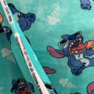 May include: Blue and white fabric featuring a repeating pattern of Stitch, a blue alien character from the Disney film Lilo & Stitch. Stitch is depicted with a mischievous expression and is holding a pink ice cream cone. The fabric has a soft, fuzzy texture.