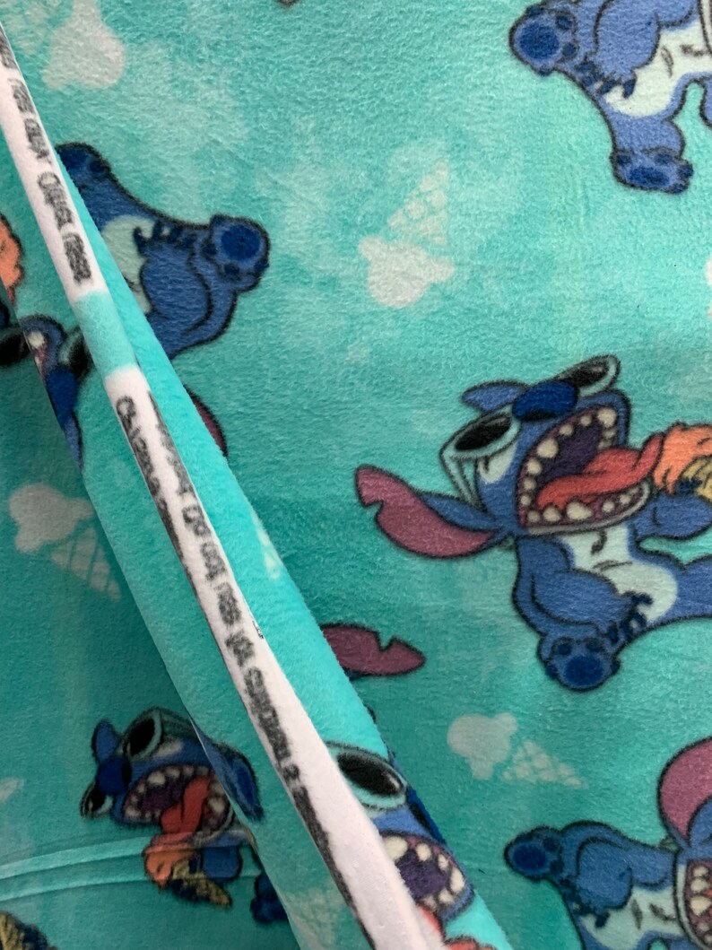 May include: Blue and white fabric featuring a repeating pattern of Stitch, a blue alien character from the Disney film Lilo & Stitch. Stitch is depicted with a mischievous expression and is holding a pink ice cream cone. The fabric has a soft, fuzzy texture.