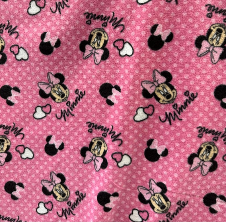 May include: Pink fabric with a white polka dot pattern featuring repeated images of Minnie Mouse with the text "Minnie" printed on the fabric.