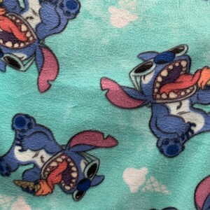 May include: A turquoise blue fabric with a repeating pattern of a blue cartoon character eating an ice cream cone. The character has large eyes, a wide open mouth, and a pink tongue sticking out. The character is wearing sunglasses.