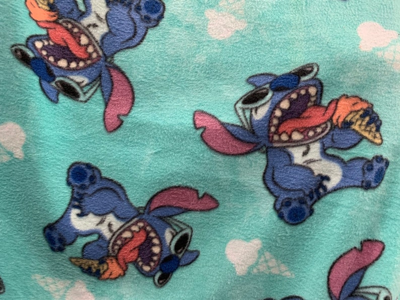 May include: A turquoise blue fabric with a repeating pattern of a blue cartoon character eating an ice cream cone. The character has large eyes, a wide open mouth, and a pink tongue sticking out. The character is wearing sunglasses.