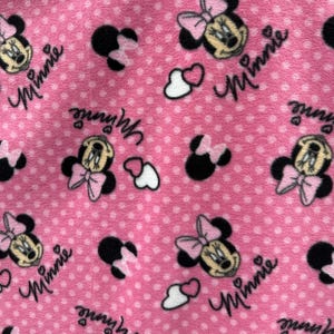 May include: Pink fabric with a repeating pattern of Minnie Mouse heads, hearts, and the word "Minnie" in a white script font.