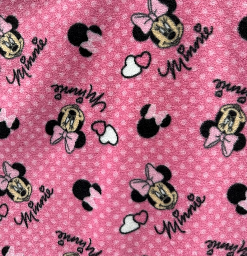 May include: Pink fabric with a repeating pattern of Minnie Mouse heads, hearts, and the word "Minnie" in a white script font.