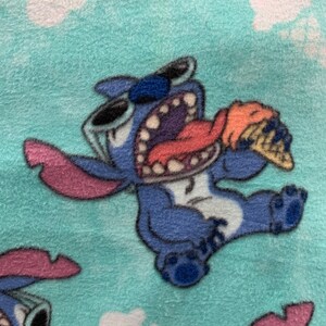 May include: Blue Stitch character with sunglasses and a pink tongue eating an orange and white ice cream cone on a turquoise background with white clouds.