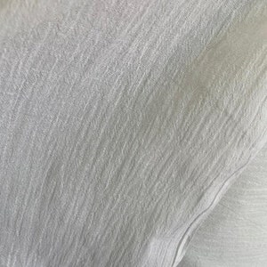 Op de afbeelding: Close-up van een witte stof met een textuur, geweven patroon. De stof heeft een zachte, licht gekreukelde uitstraling.