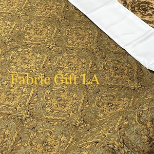 Puede incluir: Primer plano de una tela con un patr&oacute;n detallado y ornamentado en tonos dorados y marrones. La tela tiene una apariencia texturizada y una secci&oacute;n blanca en la esquina superior derecha. El texto "Fabric Gift LA" es visible en la esquina inferior izquierda.