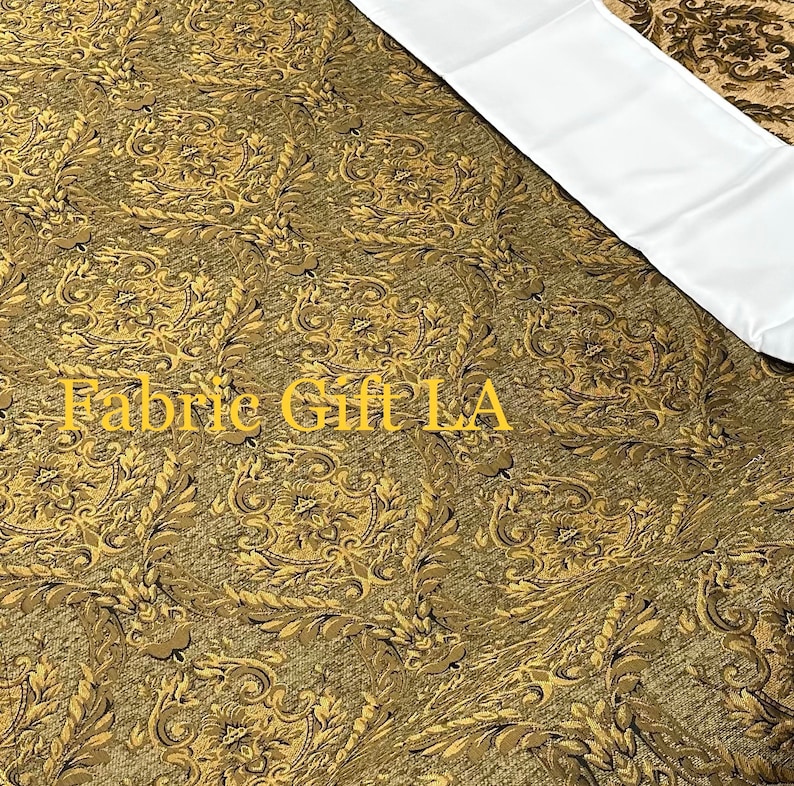 Puede incluir: Primer plano de una tela con un patr&oacute;n detallado y ornamentado en tonos dorados y marrones. La tela tiene una apariencia texturizada y una secci&oacute;n blanca en la esquina superior derecha. El texto "Fabric Gift LA" es visible en la esquina inferior izquierda.