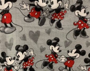 Op de afbeelding: Een grijze stof met een herhalend patroon van Mickey Mouse en Minnie Mouse die hand in hand lopen. De achtergrond is lichtgrijs met kleine grijze hartjes.