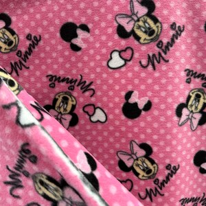 May include: Pink fleece fabric with a repeating pattern of Minnie Mouse heads, hearts, and polka dots. The fabric has the word "Minnie" written on it.