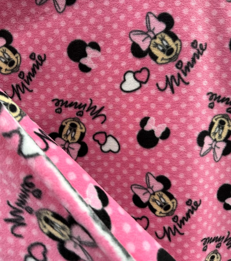 May include: Pink fleece fabric with a repeating pattern of Minnie Mouse heads, hearts, and polka dots. The fabric has the word "Minnie" written on it.