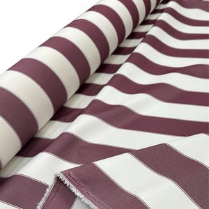 May include: A roll of fabric with wide, alternating stripes of burgundy and white. The fabric is partially unrolled, revealing the pattern and the raw edge of the material. The stripes are evenly spaced and create a bold, classic design.