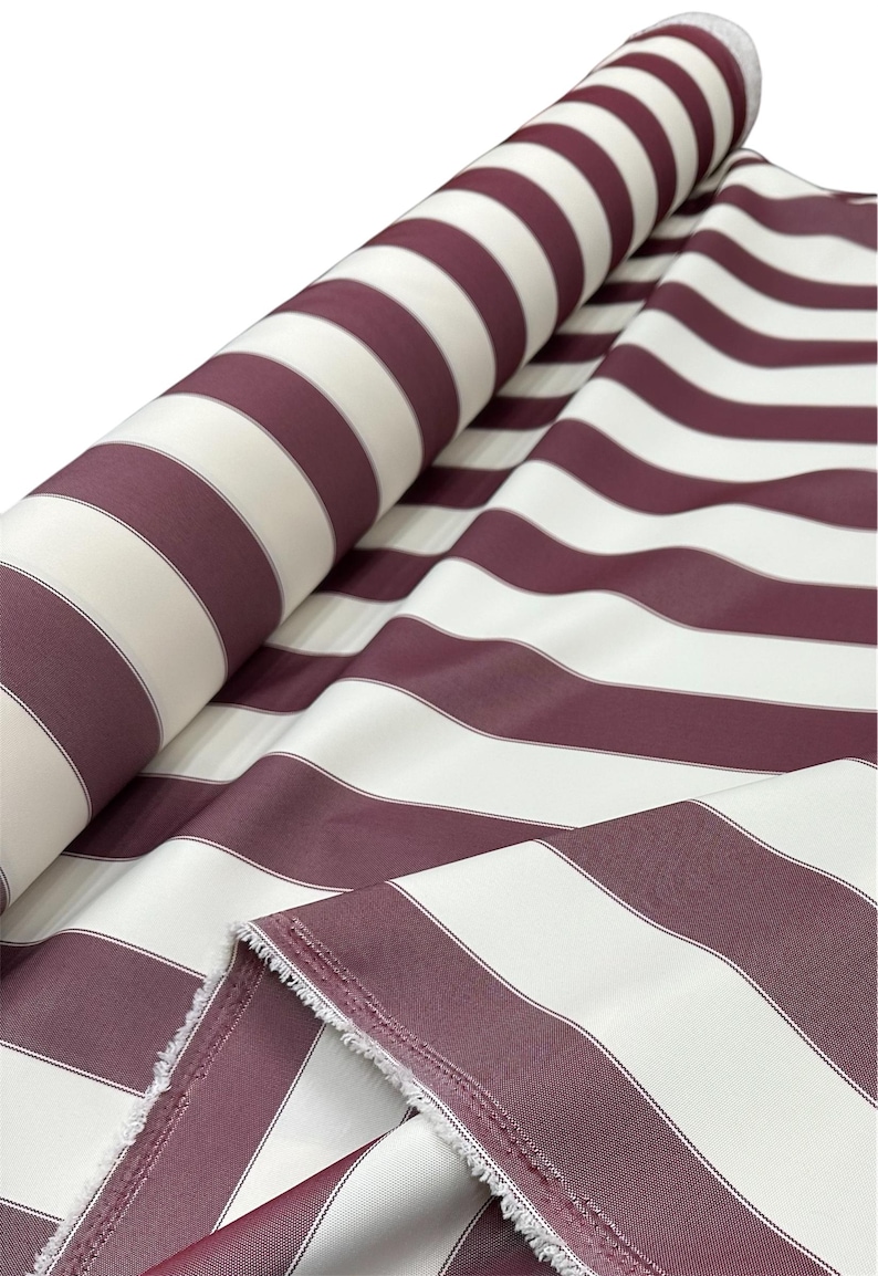 May include: A roll of fabric with wide, alternating stripes of burgundy and white. The fabric is partially unrolled, revealing the pattern and the raw edge of the material. The stripes are evenly spaced and create a bold, classic design.
