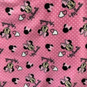 May include: Pink fabric with a repeating pattern of Minnie Mouse heads, white hearts, and black polka dots. The text "Minnie" is repeated throughout the fabric.