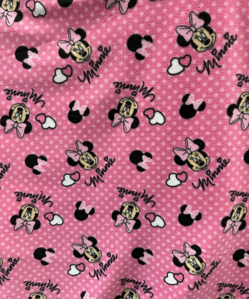 May include: Pink fabric with a repeating pattern of Minnie Mouse heads, white hearts, and black polka dots. The text "Minnie" is repeated throughout the fabric.