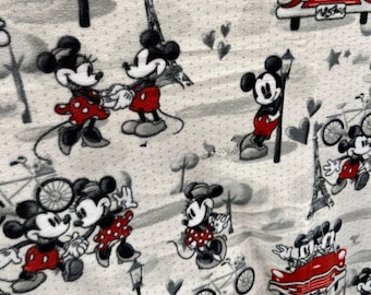 Op de afbeelding: Een stof met een herhalend patroon van Mickey en Minnie Mouse in Parijs. Het ontwerp omvat de Eiffeltoren, fietsen, rode auto's en harten. Mickey en Minnie worden in verschillende poses afgebeeld, met een kleurenschema van zwart, rood en wit.