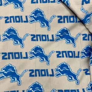 May include: A tan fabric with a repeating pattern of blue Detroit Lions logos and the word "LIONS" in blue. The logos are of a lion in a running pose.