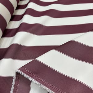 May include: Close-up of a fabric roll featuring wide, horizontal stripes. Alternating bands of burgundy and white create a bold pattern. The fabric has a slight sheen, and the edges show a raw, unfinished texture. Ideal for upholstery or drapery.