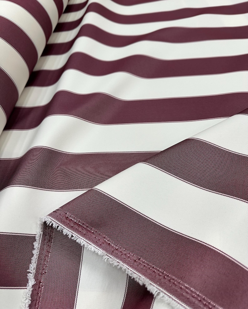 May include: Close-up of a fabric roll featuring wide, horizontal stripes. Alternating bands of burgundy and white create a bold pattern. The fabric has a slight sheen, and the edges show a raw, unfinished texture. Ideal for upholstery or drapery.