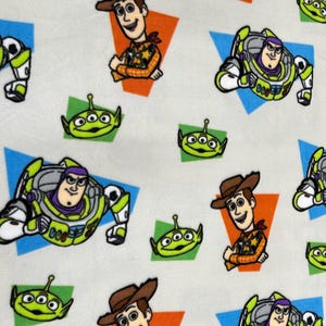 May include: Fabric with a repeating pattern of Toy Story characters. The design includes Buzz Lightyear in a green and white spacesuit, Woody the cowboy, and the green three-eyed aliens. The characters are set against colourful geometric shapes.