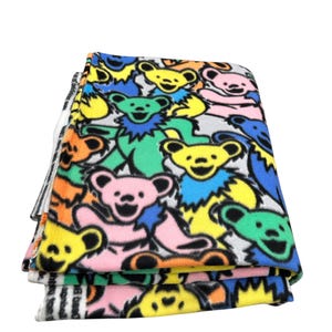 Puede incluir: Manta de forro polar plegada con un patr&oacute;n colorido de osos bailarines en amarillo, rosa, verde y azul. La manta tiene un fondo blanco y un borde a rayas blanco y negro.