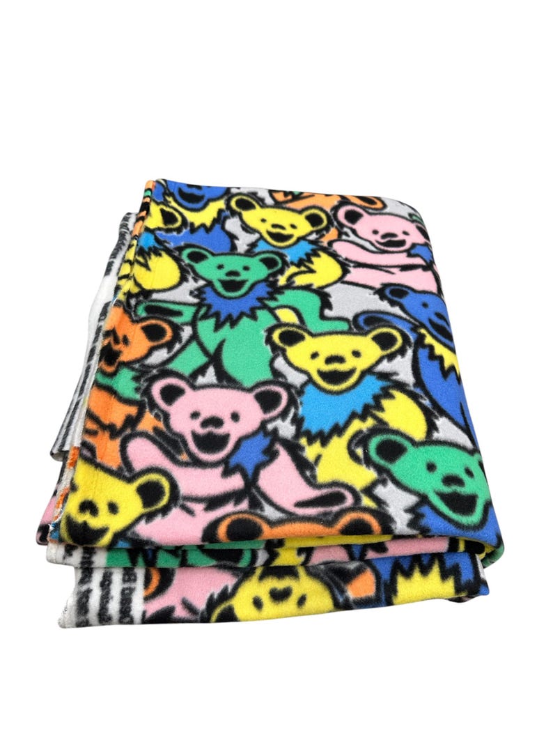Puede incluir: Manta de forro polar plegada con un patr&oacute;n colorido de osos bailarines en amarillo, rosa, verde y azul. La manta tiene un fondo blanco y un borde a rayas blanco y negro.