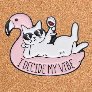 May include: White cat wearing sunglasses and holding a glass of wine, relaxing on a pink flamingo pool float. The float has the text "I decide my vibe" written on it.