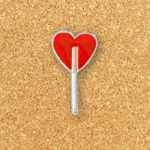 Puede incluir: Una piruleta roja en forma de coraz&oacute;n en un palo blanco, fijada a un tablero de corcho.