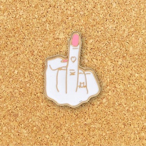 May include: White enamel pin of a middle finger with gold trim and pink nail polish. The finger has a small heart tattoo and a ring on the ring finger.